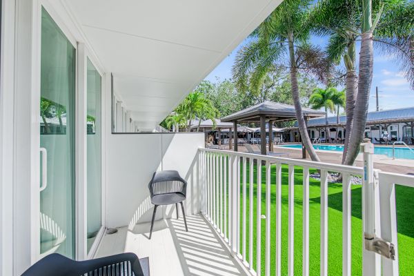 A balcony with two chairs overlooks a garden area with palm trees and a pool, featuring lounge spaces and a clear blue sky.