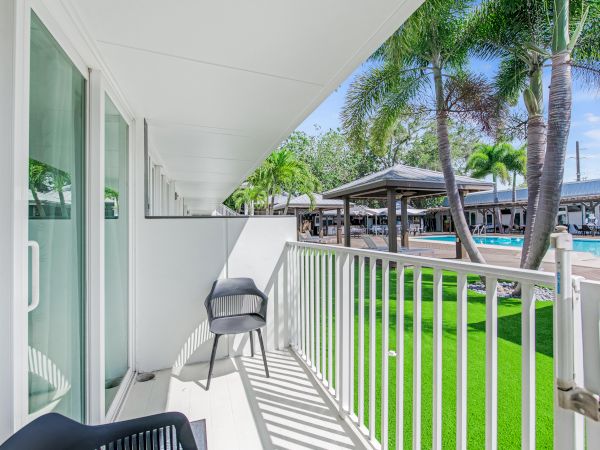 A balcony with two chairs overlooks a garden area with palm trees and a pool, featuring lounge spaces and a clear blue sky.