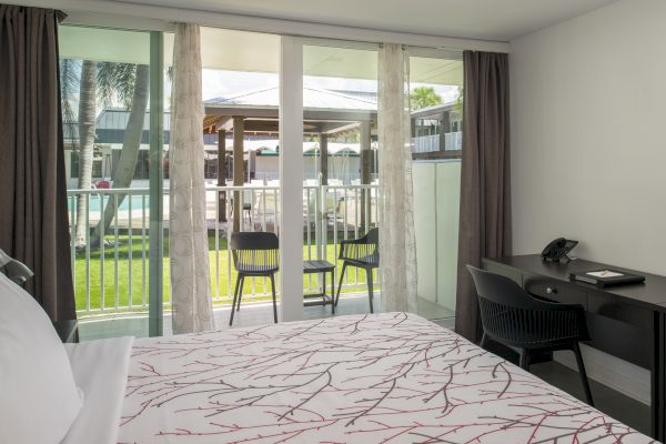 A modern bedroom features a neatly made bed, desk, and chairs on a balcony overlooking a garden and pool area, framed by glass sliding doors.