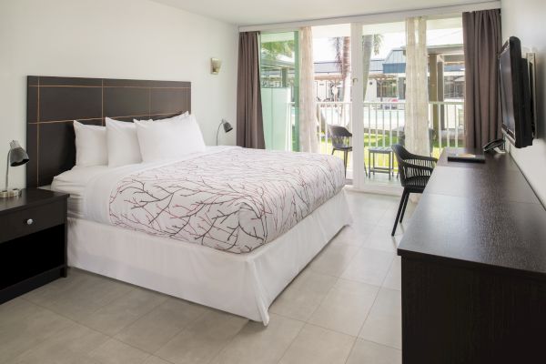 A modern hotel room with a large bed, TV, desk, and chairs outside on the balcony, adorned with neutral decor and large windows.