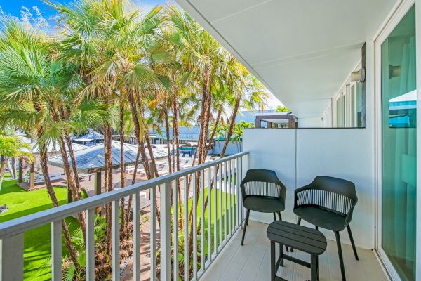 A cozy balcony with two chairs and a small table, overlooking a garden full of palm trees on a sunny day.