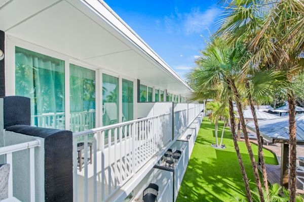 A modern building with white balconies and large windows, surrounded by green palm trees and a bright blue sky.