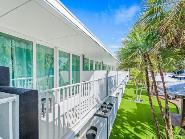 A modern building with white balconies and large windows, surrounded by green palm trees and a bright blue sky.