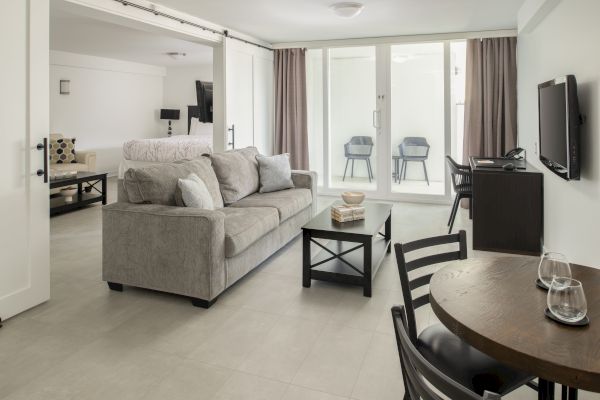 A modern living room features a gray sofa, a coffee table, a TV, a small dining table, and large windows with curtains leading to a balcony.