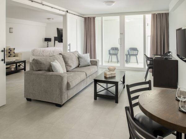 A modern living room features a gray sofa, a coffee table, a TV, a small dining table, and large windows with curtains leading to a balcony.