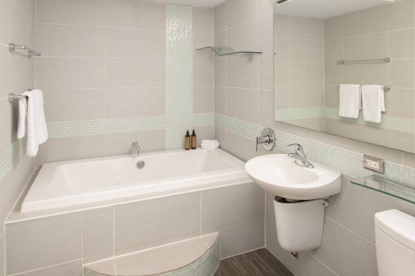 A modern bathroom with a bathtub, sink, and toilet; towels, soap dispensers, and a large mirror on the wall.