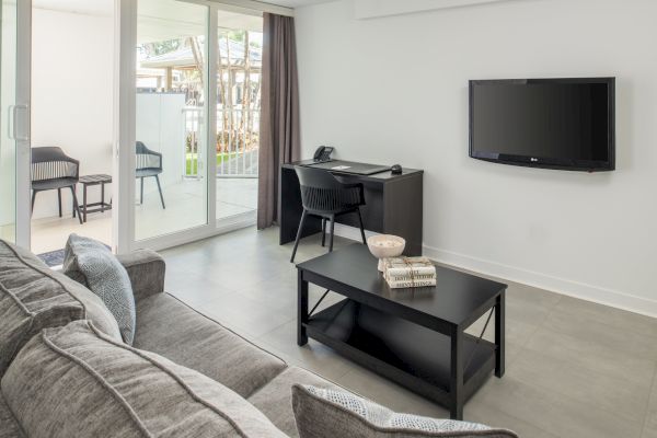 A modern living room with a gray sofa, coffee table, wall-mounted TV, and desk by a glass door leading to an outdoor seating area.