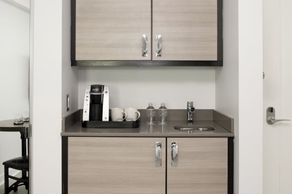 A compact kitchenette with light-colored cabinets, a coffee machine, mugs, water bottles, and a small sink, clearly organized in a modern setting.