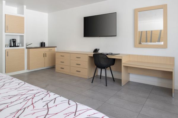 A tidy hotel room features a desk, chair, TV, mirror, and a counter with a coffee maker against a wall with light wood cabinets.