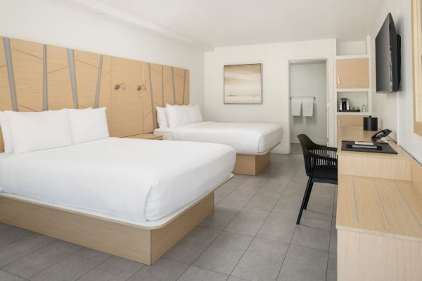 A hotel room with two double beds, a wall-mounted TV, a desk with a chair, a minimalist decor, and an open door revealing towels in a bathroom.