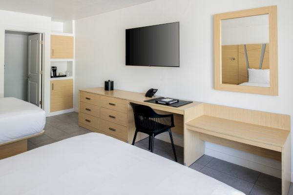 The image shows a modern hotel room with two beds, a TV, a desk with a chair, a mirror, and a cabinet with a coffee maker.