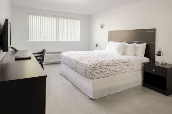 This image shows a modern, minimalist bedroom with a bed, nightstands, a TV on a dresser, a chair by the window, and contemporary decor.