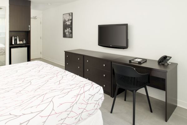 The image shows a hotel room with a bed, a TV mounted on the wall, a desk with a chair, a phone, and an open closet with amenities.