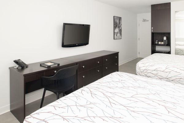 The image shows a hotel room with two beds, a wall-mounted TV, a desk with a chair and phone, a dresser, and a black and white photo on the wall.