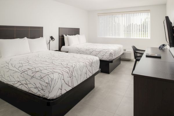 The image shows a tidy hotel room with two double beds, a flat-screen TV, a desk with a chair, and a window with vertical blinds.