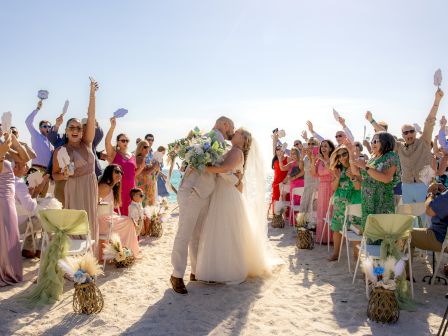 A newly married couple is kissing on a beach, surrounded by cheering, colorfully dressed guests, raising their hands in celebration.