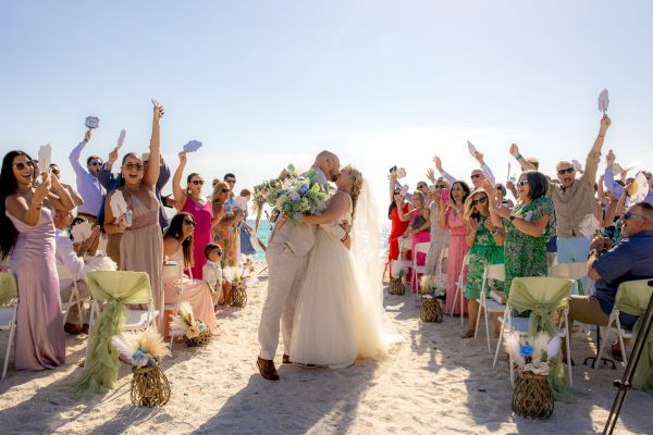 A newly married couple is kissing on a beach, surrounded by cheering, colorfully dressed guests, raising their hands in celebration.