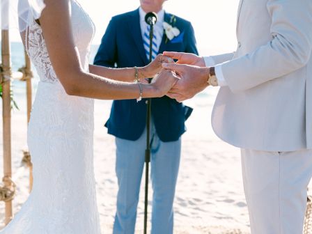 A bride and groom hold hands during their wedding ceremony on a beach, with an officiant standing behind them speaking into a microphone.