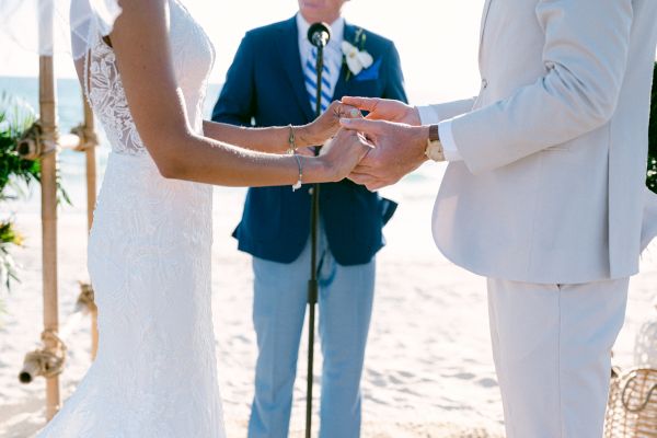 A bride and groom hold hands during their wedding ceremony on a beach, with an officiant standing behind them speaking into a microphone.