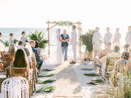 A beach wedding ceremony with a couple exchanging vows under a wooden arch, attended by guests seated on either side of the aisle.