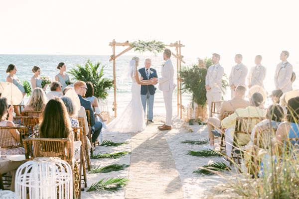 A beach wedding ceremony with a couple exchanging vows under a wooden arch, attended by guests seated on either side of the aisle.