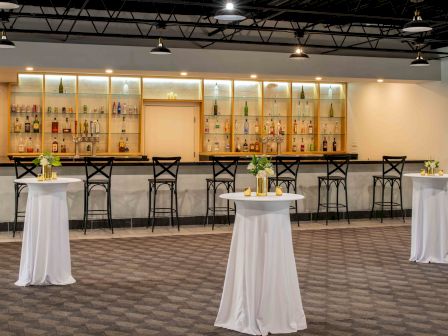 The image shows a modern bar with high shelves of bottles, several barstools, and three standing tables draped in white cloths adorned with flowers.