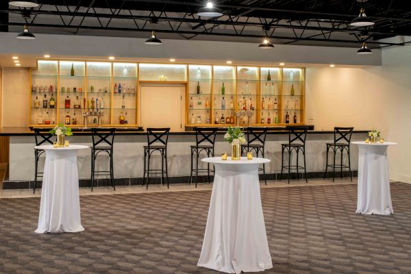 The image shows a modern bar with high shelves of bottles, several barstools, and three standing tables draped in white cloths adorned with flowers.