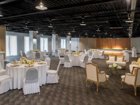 A decorated banquet hall with round tables covered with white cloth and surrounded by chairs, set for an event or gathering, and lounge seating.