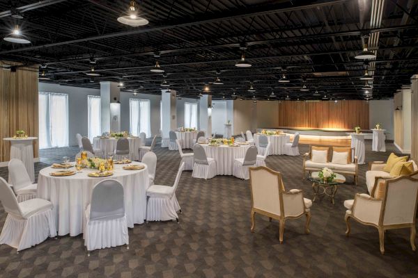 A decorated banquet hall with round tables covered with white cloth and surrounded by chairs, set for an event or gathering, and lounge seating.