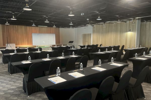 This image depicts a conference room setup with black-draped tables, chairs, water bottles, notebooks, and a projector screen at the front.