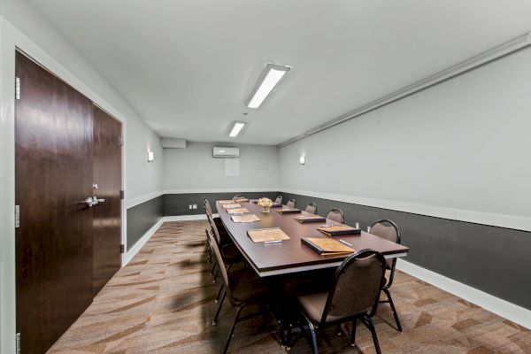 A conference room with a long table, chairs, notebooks, and pens, illuminated by ceiling lights and featuring a patterned carpet.