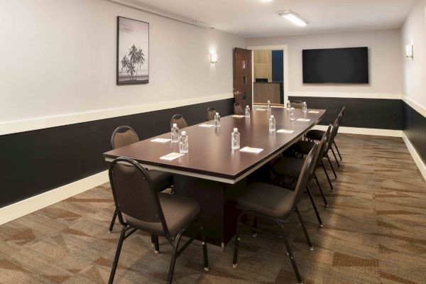 A conference room with a rectangular table, eight chairs, water bottles, notepads, a painting, and a TV screen on the wall are visible.