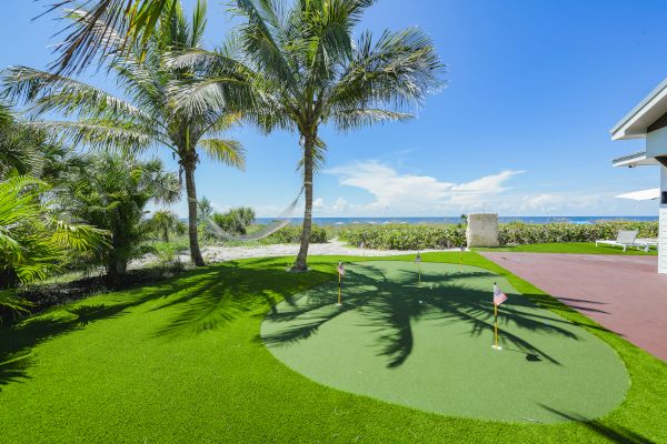 A tropical backyard features a putting green, palm trees, a hammock, and a view of the ocean under a clear blue sky, next to a house.