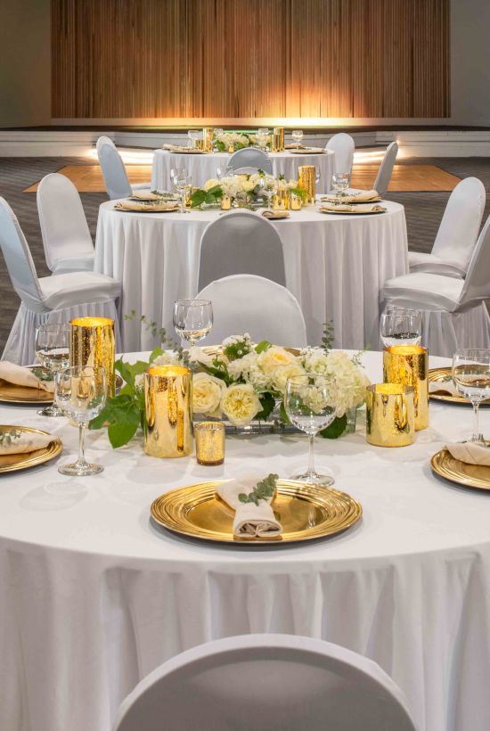 The image shows an elegantly set indoor event space with round tables, white chairs, flower centerpieces, and golden decorative elements.