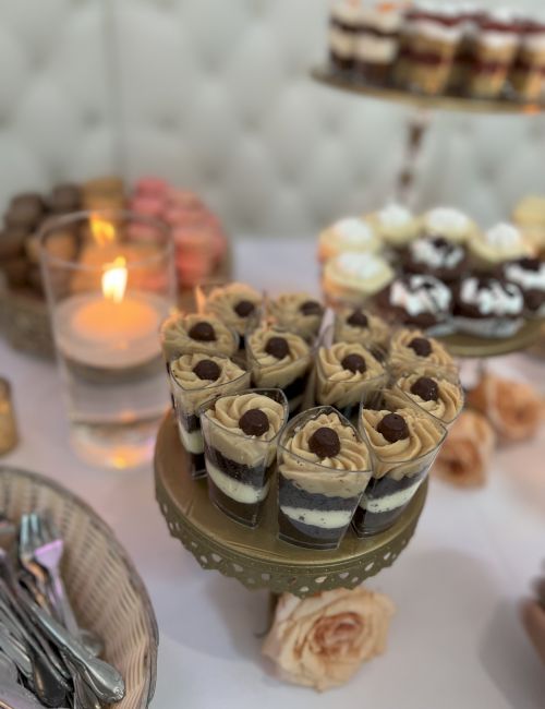 The image shows a dessert table with various sweets, including layered parfaits in small cups, cupcakes, and a lit candle in a glass holder.