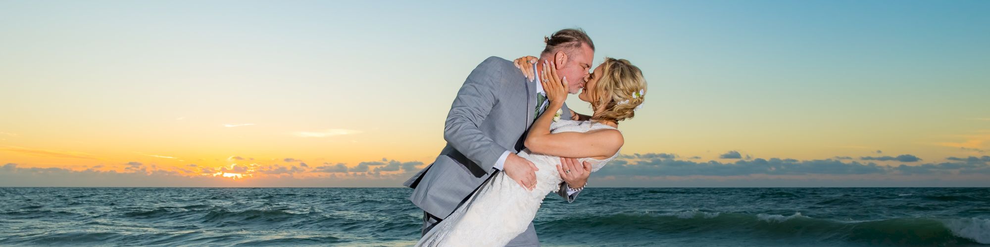 A couple in wedding attire shares a romantic kiss on a beach at sunset, with ocean waves in the background.