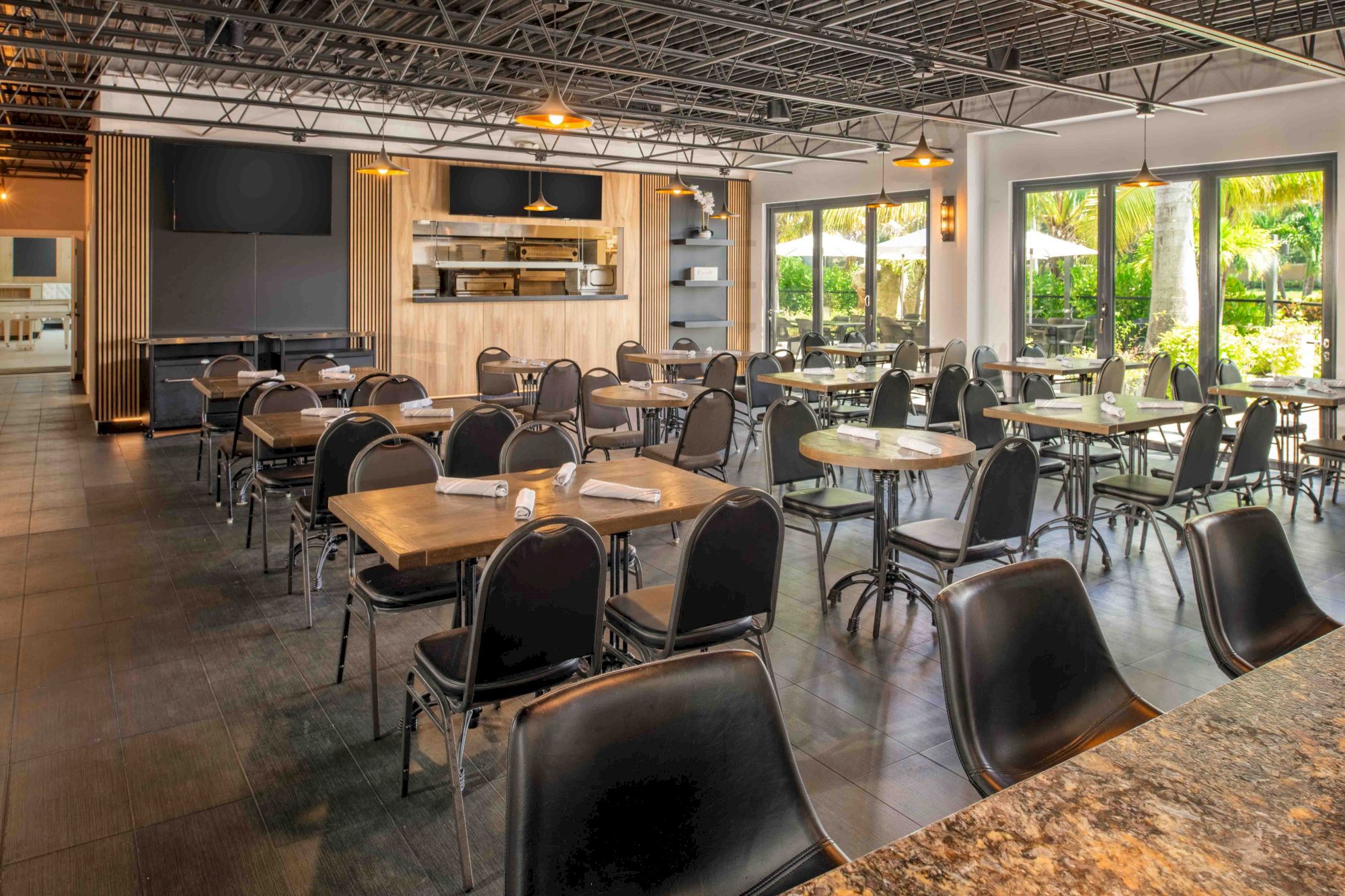 The image shows an empty, well-lit restaurant with modern decor, featuring several tables and chairs, large windows, and a view of greenery outside.