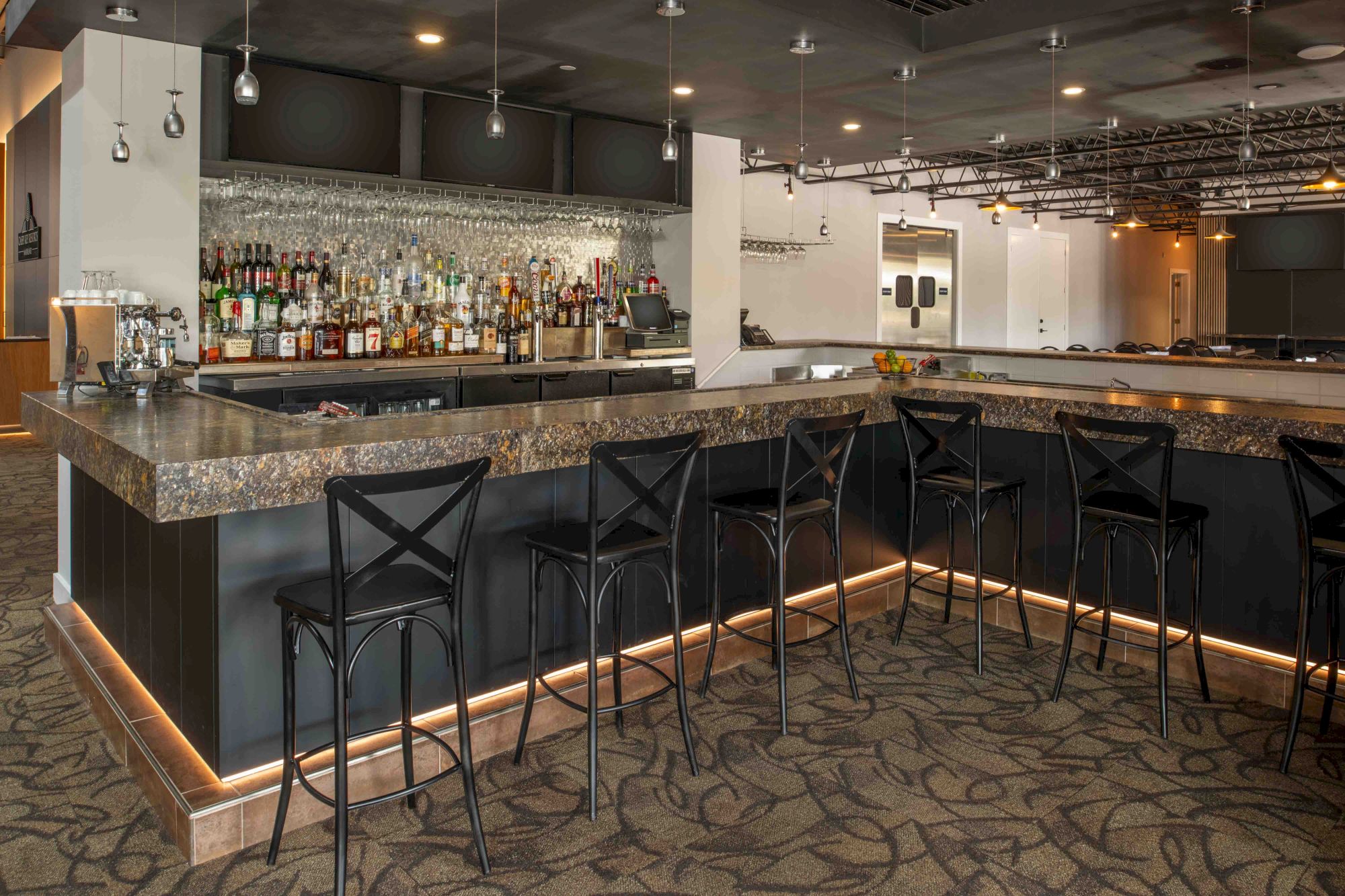 The image shows a modern bar with a well-stocked shelf of liquor bottles, a marble countertop, and black barstools. Ending the sentence.