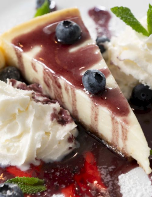 The image shows a slice of cheesecake with blueberry sauce, whipped cream, blueberries, a scoop of vanilla ice cream, and mint leaves on a plate.