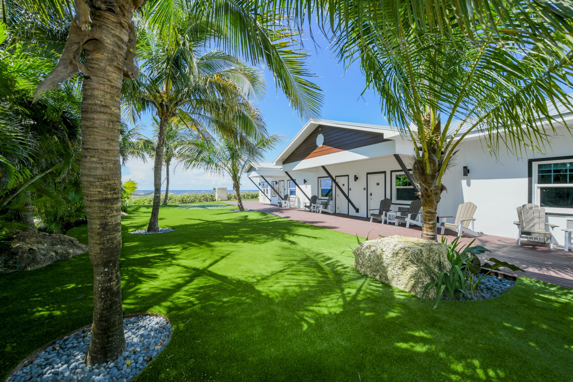 A modern, white bungalow with a patio, surrounded by lush green grass, palm trees, and outdoor seating is shown in the image.