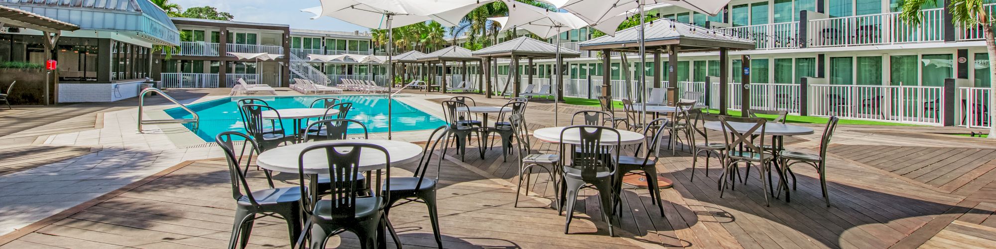 An outdoor patio with tables, chairs, umbrellas, and string lights next to a pool area, surrounded by modern-looking buildings against a clear sky.