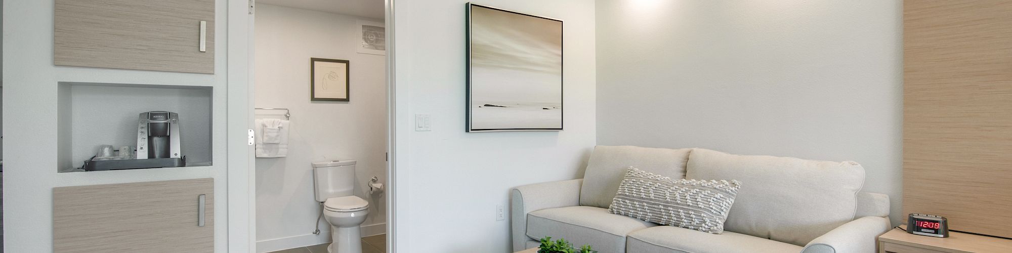 The image shows a modern living space with a couch, coffee table, small bathroom, and cabinets on tiled flooring.