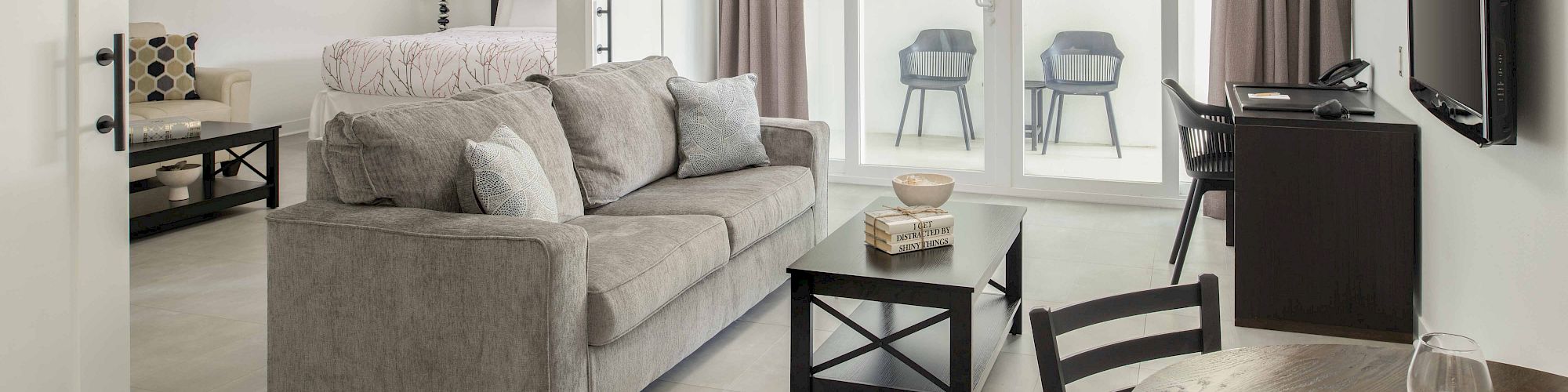 This image shows a modern living area with a sofa, coffee table, TV, and a small dining table with chairs, leading to a balcony with seating.