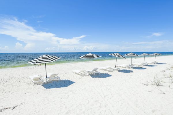 A sandy beach with several black-and-white striped umbrellas and white lounge chairs facing the clear blue ocean under a sunny sky with clouds.