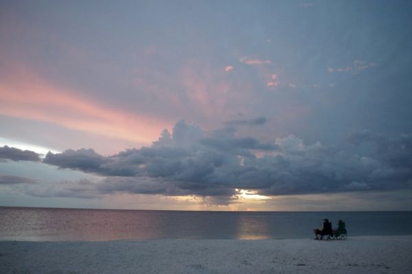 A serene beach scene at sunset with two people sitting on chairs near the water, under a sky with clouds in pink and blue hues.