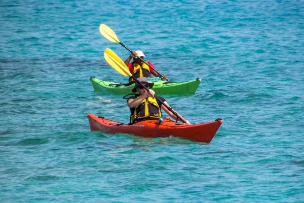 Two people are kayaking in clear blue waters, each in a kayak with yellow paddles, one in a red kayak and the other in a green kayak.