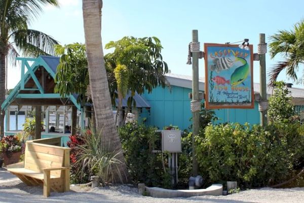 A tropical-themed restaurant or shop with palm trees and vibrant signage, featuring outdoor seating and lush greenery in a sunny environment.