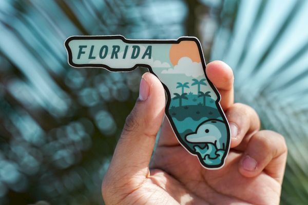 A hand holds a Florida-shaped sticker featuring a dolphin, ocean, palm trees, and a setting sun, with blurred palm leaves in the background.