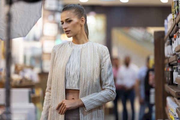 A woman in a chic outfit is standing indoors with blurred shelves and people in the background, seemingly in a photo shoot setting.