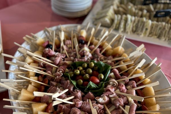 A platter of skewered appetizers including meat and cheese is in the foreground, with plates and more food items in the background on a table.